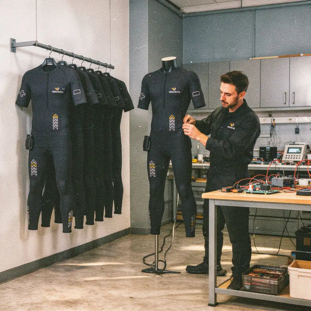 A technician fitting a Visionbody EMS suit to a mannequin beside an electronics workbench in Germany