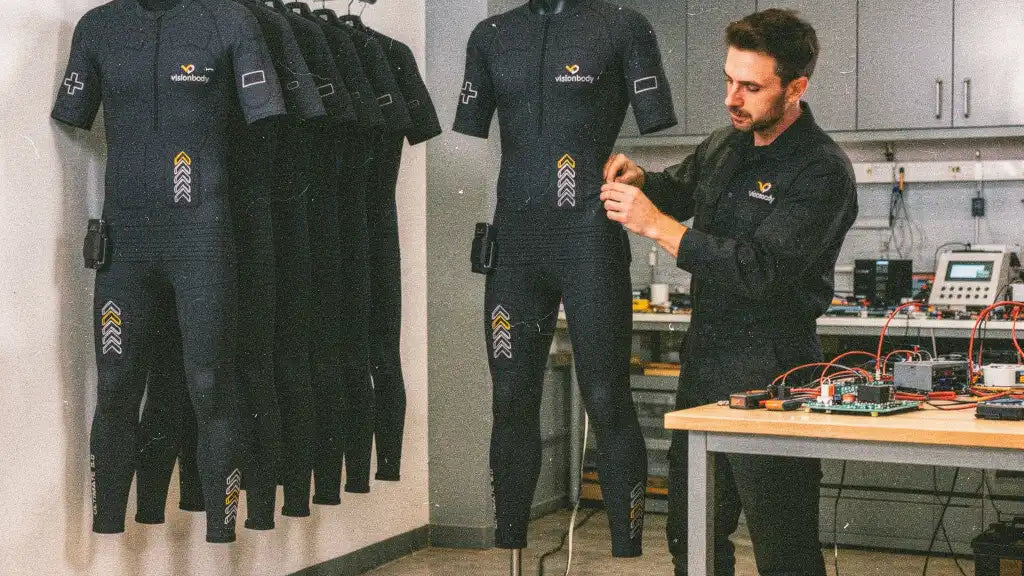 A technician fitting a Visionbody EMS suit to a mannequin beside an electronics workbench in Germany